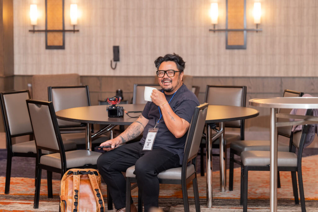 A man with photography equipment sitting at a round table holding up a coffee mug and smiling
