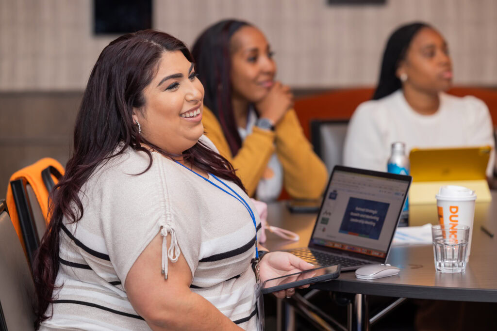 A woman at a round table watching a conference speaker and smiling