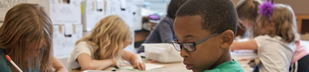 seated amongst his classmates, this photograph depicts a young Black schoolboy who was in the process of drawing with a pencil on a piece of white paper.